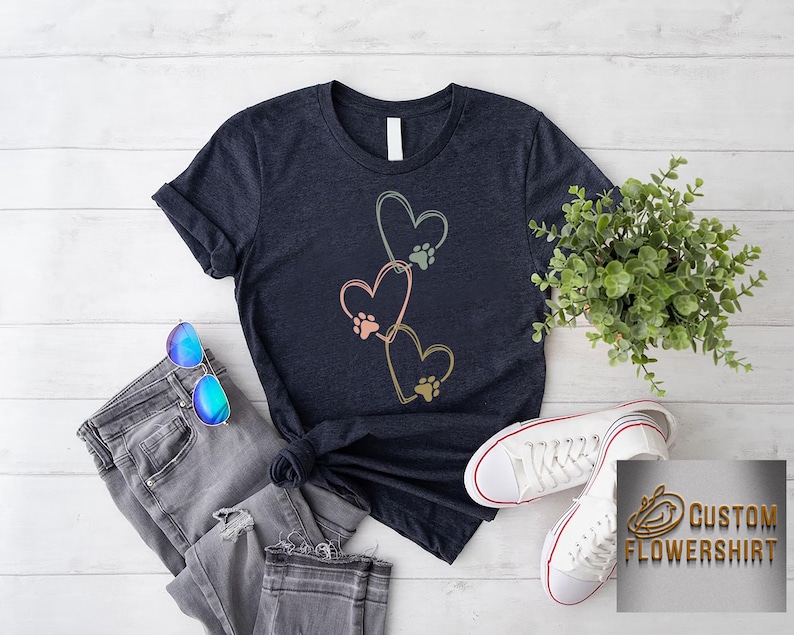 Pode incluir: Camiseta azul marinho com tr&ecirc;s cora&ccedil;&otilde;es e pegadas em rosa, verde e dourado. A camiseta &eacute; combinada com jeans cinza, &oacute;culos de sol azuis e t&ecirc;nis brancos. Uma pequena planta e um logotipo tamb&eacute;m s&atilde;o vis&iacute;veis.