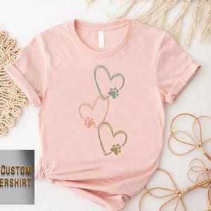 Pode incluir: Camiseta rosa claro com tr&ecirc;s cora&ccedil;&otilde;es conectados, cada um com uma pegada de pata. Os cora&ccedil;&otilde;es s&atilde;o delineados em verde. O texto "CUSTOM FLOWERSHIRT" &eacute; vis&iacute;vel no canto inferior esquerdo.