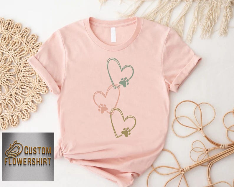 Pode incluir: Camiseta rosa claro com tr&ecirc;s cora&ccedil;&otilde;es conectados, cada um com uma pegada de pata. Os cora&ccedil;&otilde;es s&atilde;o delineados em verde. O texto "CUSTOM FLOWERSHIRT" &eacute; vis&iacute;vel no canto inferior esquerdo.