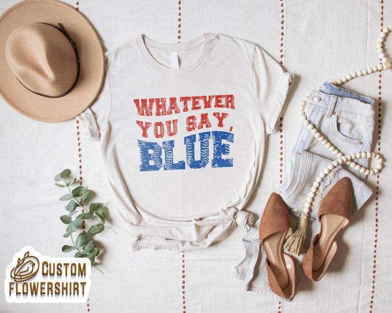 Może przedstawiać: Kremowy t-shirt z napisem "WHATEVER YOU SAY, BLUE" w czerwonych i niebieskich literach. Koszulka jest stylizowana z jasnymi dżinsami, brązowymi balerinami, naszyjnikiem z koralik&oacute;w i beżowym kapeluszem.
