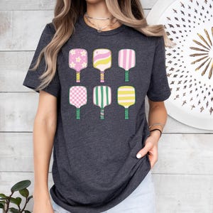 May include: Dark gray t-shirt featuring a colorful graphic of six pickleball paddles. The paddles have various patterns, including flowers, stripes, and checks, in shades of pink, yellow, and green. The shirt is a casual, everyday wear item.
