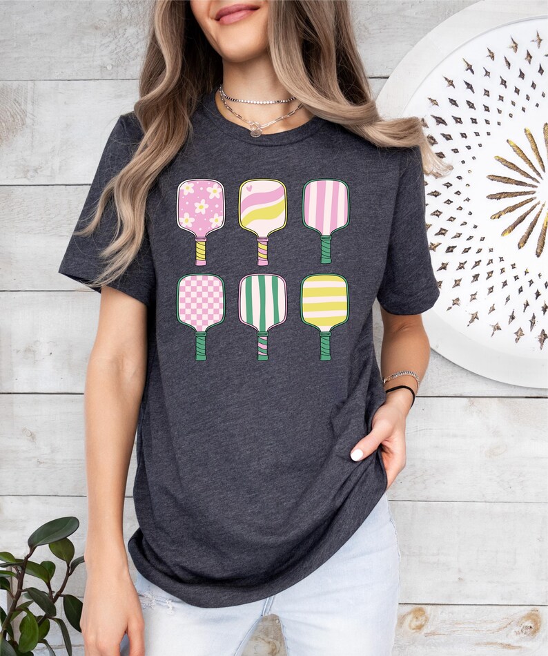 May include: Dark gray t-shirt featuring a colorful graphic of six pickleball paddles. The paddles have various patterns, including flowers, stripes, and checks, in shades of pink, yellow, and green. The shirt is a casual, everyday wear item.