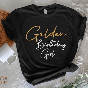 May include: Black t-shirt with the text "Golden Birthday Girl" in gold and white lettering.