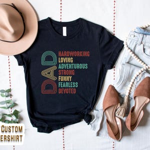 May include: Black t-shirt with a retro-style graphic that says "DAD" with the letters made up of colorful arcs. The text below the letters says "Hardworking, Loving, Adventurous, Strong, Funny, Fearless, Devoted".