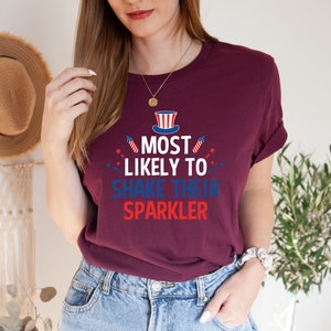 Op de afbeelding: Een maroon t-shirt met een witte, rode en blauwe afbeelding met de tekst "Most Likely to Shake Their Sparkler". De afbeelding bevat een kleine Amerikaanse vlag en vuurwerk.