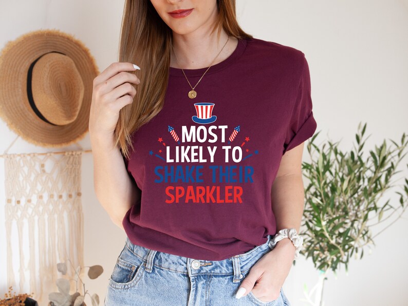 Op de afbeelding: Een maroon t-shirt met een witte, rode en blauwe afbeelding met de tekst "Most Likely to Shake Their Sparkler". De afbeelding bevat een kleine Amerikaanse vlag en vuurwerk.