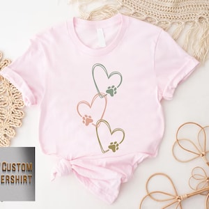 Pode incluir: Camiseta rosa claro com tr&ecirc;s cora&ccedil;&otilde;es conectados, cada um com uma pegada de pata. Os contornos dos cora&ccedil;&otilde;es s&atilde;o verdes, rosa e dourado. O texto "CUSTOM FLOWERSHIRT" &eacute; vis&iacute;vel no canto inferior esquerdo.