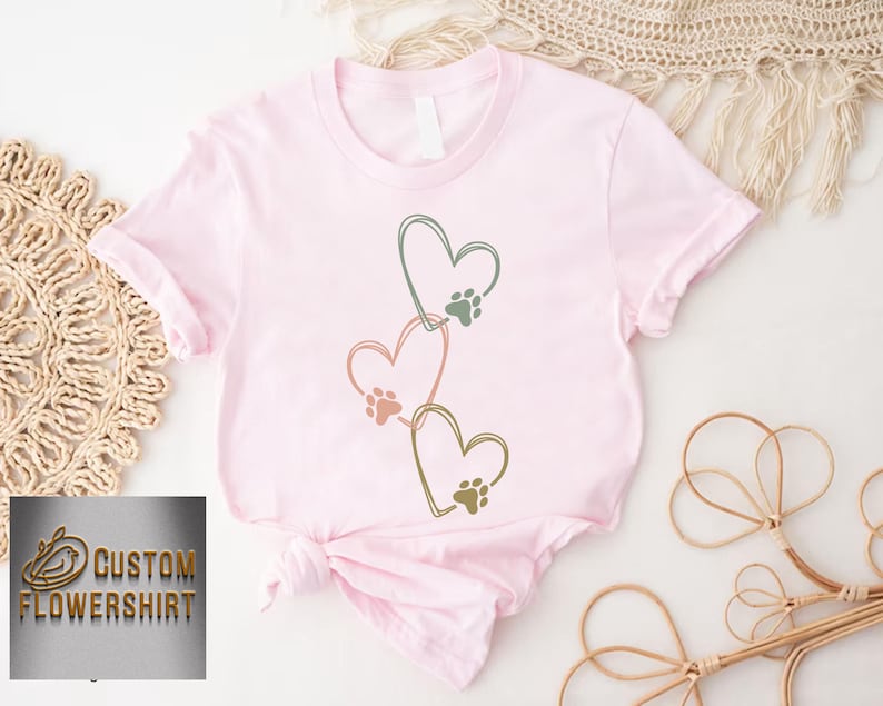 Pode incluir: Camiseta rosa claro com tr&ecirc;s cora&ccedil;&otilde;es conectados, cada um com uma pegada de pata. Os contornos dos cora&ccedil;&otilde;es s&atilde;o verdes, rosa e dourado. O texto "CUSTOM FLOWERSHIRT" &eacute; vis&iacute;vel no canto inferior esquerdo.