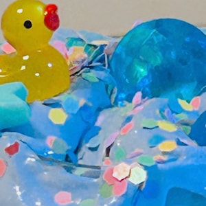May include: A yellow rubber ducky with a red beak and blue eyes is partially submerged in a blue slime with colorful confetti. A blue glass orb is also partially submerged in the slime.