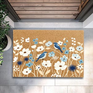 May include: A rectangular doormat with a natural coir fiber background. The mat features a floral design with white and light blue flowers, and two blue birds. The mat is 24 inches wide and 36 inches long.