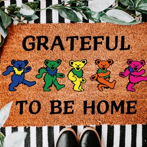 May include: A brown doormat with the words "GRATEFUL TO BE HOME" in black. Five colorful dancing bear figures are below the text. The doormat is on a black and white striped surface.