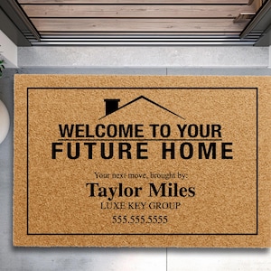 May include: A rectangular doormat with a black border and the text "WELCOME TO YOUR FUTURE HOME" in black. Below the text is "Your next move, brought by: Taylor Miles LUXE KEY GROUP 555.555.5555". The mat is made of a light brown material.