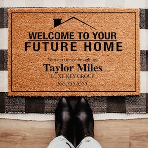 May include: A rectangular doormat with the text "WELCOME TO YOUR FUTURE HOME" in black. The mat is brown with a black border and sits on a black and white checkered rug. The text "Taylor Miles" and a phone number are also visible.