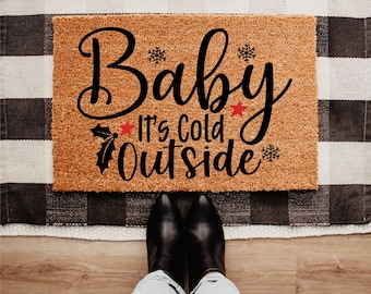 May include: A rectangular doormat with the text "Baby It's Cold Outside" in black script. The mat is brown with a textured surface, and the text is decorated with snowflakes, a red star, and holly. The mat is placed on a black and white checkered rug.