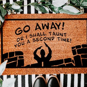 May include: A rectangular doormat with the text "GO AWAY! OR I SHALL TAUNT YOU A SECOND TIME!" in black. The mat has a brown background with a black border and a silhouette of a person and castle. The mat is on a black and white striped surface.