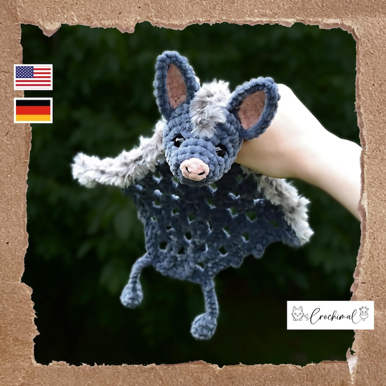 May include: A handmade crochet bat in shades of blue and gray. The bat has large ears, a pink nose, and small black eyes. The wings are a darker blue with a textured pattern. The bat is held against a green background.