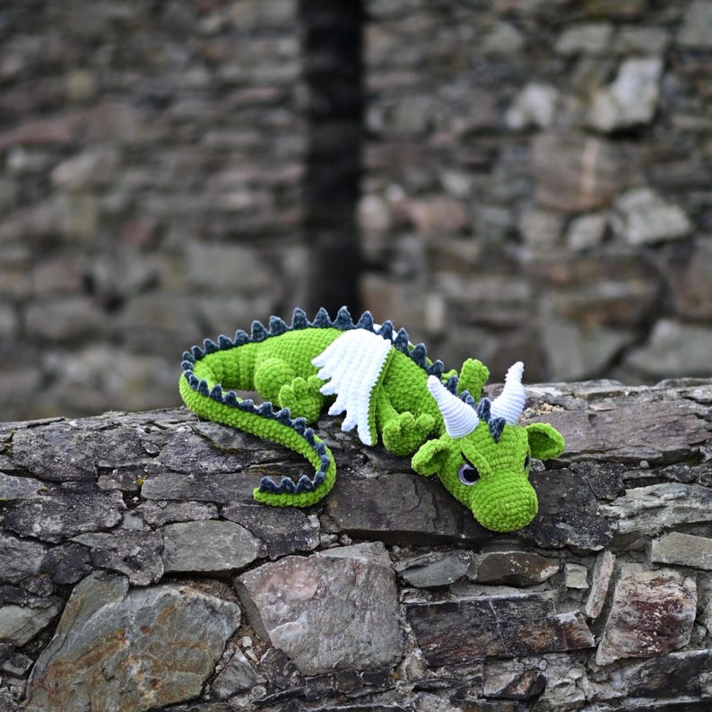 K&ouml;nnte beinhalten: Ein handgefertigter gr&uuml;ner H&auml;kchendrache mit wei&szlig;en Fl&uuml;geln und blau gef&auml;rbten H&ouml;rnern liegt auf einer Steinmauer. Der Drache hat einen langen, geschwungenen Schwanz und einen detaillierten, strukturierten K&ouml;rper. Der Hintergrund ist eine verschwommene Steinmauer.