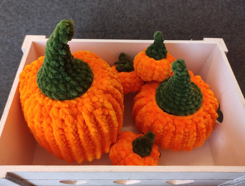 May include: Five crocheted pumpkins in a white wooden crate. The pumpkins are orange with green stems.