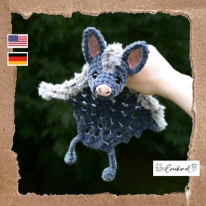 Pode incluir: Um brinquedo de morcego de crochê feito à mão em tons de azul e cinza. O morcego tem orelhas grandes, um nariz rosa e um corpo cinza fofo. As asas são de um azul mais escuro com um padrão texturizado. O brinquedo é segurado contra um fundo verde.