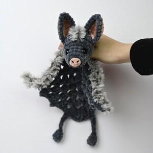 May include: A handmade crochet bat toy in shades of gray and black. The bat has large ears, a pink nose, and a fluffy gray and black body. The wings are a dark gray with a textured pattern. The bat is held against a white background.