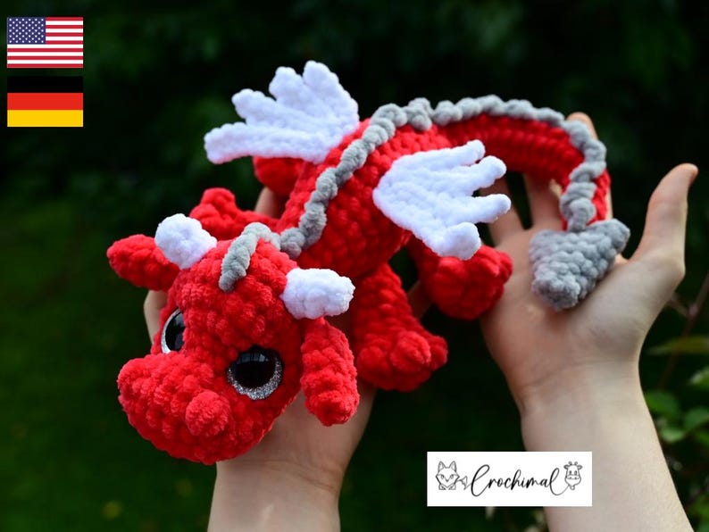 May include: A red crocheted dragon plush toy featuring white wings, white horns, and a gray spine ridge that extends to a gray tail tip. The dragon has large, round, black eyes with silver glitter accents and a textured, soft yarn construction.