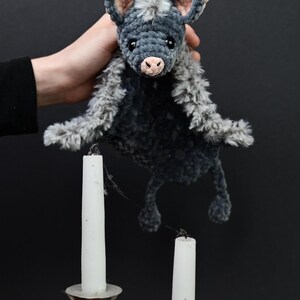 May include: A handmade plush bat in shades of gray, with a pink nose and inner ears. The bat is held in front of two white candles in silver candle holders. The plush toy has a soft, textured appearance.