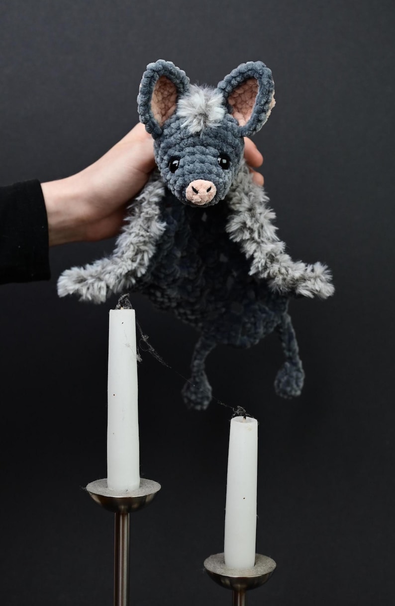 May include: A handmade plush bat in shades of gray, with a pink nose and inner ears. The bat is held in front of two white candles in silver candle holders. The plush toy has a soft, textured appearance.