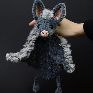 May include: A handmade plush bat toy in shades of gray and blue. The bat has large, upright ears with pink inner ears, a pink nose, and small black eyes. The body is made of soft, textured fabric, with a fluffy, gray trim around the wings.