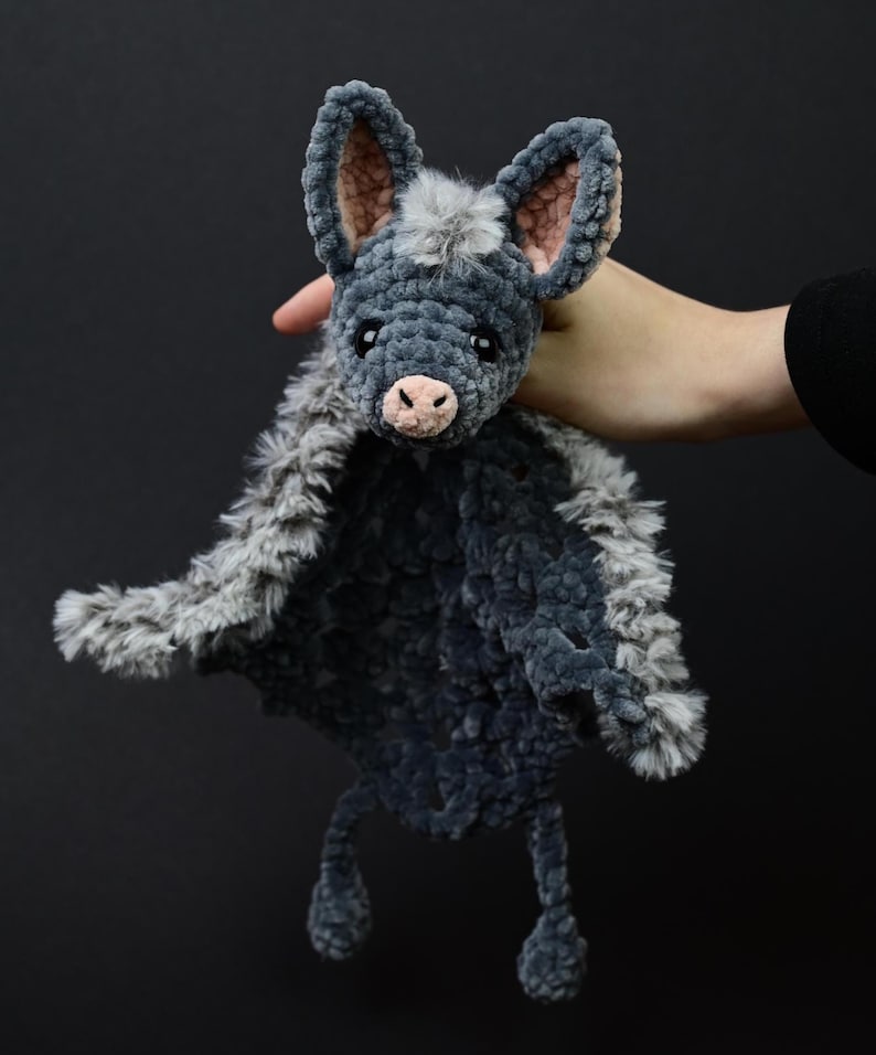 May include: A handmade plush bat toy in shades of gray and blue. The bat has large, upright ears with pink inner ears, a pink nose, and small black eyes. The body is made of soft, textured fabric, with a fluffy, gray trim around the wings.