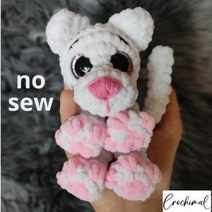 Op de afbeelding: Een wit gehaakt knuffeldier met roze accenten, waaronder de neus en poten. Het speelgoed heeft grote, glinsterende ogen en een zachte textuur. De tekst "no sew" is zichtbaar op de afbeelding.