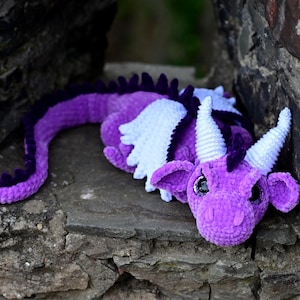 K&ouml;nnte beinhalten: Ein handgefertigter H&auml;keldraken in Lilat&ouml;nen, mit wei&szlig;en Fl&uuml;geln und H&ouml;rnern. Der Drache liegt auf einer Steinoberfl&auml;che und zeigt sein detailliertes Design. Der Schwanz des Drachen ist in einem dunkleren Lilaton gehalten.