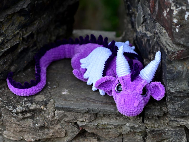 K&ouml;nnte beinhalten: Ein handgefertigter H&auml;keldraken in Lilat&ouml;nen, mit wei&szlig;en Fl&uuml;geln und H&ouml;rnern. Der Drache liegt auf einer Steinoberfl&auml;che und zeigt sein detailliertes Design. Der Schwanz des Drachen ist in einem dunkleren Lilaton gehalten.