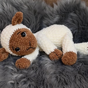 May include: A crocheted plush toy lamb with brown and white fur. The lamb is lying on a grey fluffy surface.