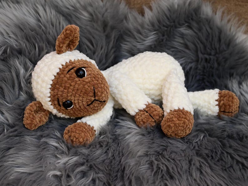 May include: A crocheted plush toy lamb with brown and white fur. The lamb is lying on a grey fluffy surface.