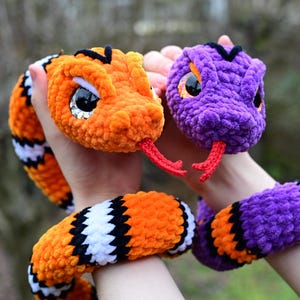 Pode incluir: Dois brinquedos de cobra de croch&ecirc;, um laranja e preto, o outro roxo e preto. Ambos t&ecirc;m olhos grandes e pretos e l&iacute;nguas vermelhas.