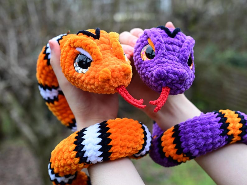 Pode incluir: Dois brinquedos de cobra de croch&ecirc;, um laranja e preto, o outro roxo e preto. Ambos t&ecirc;m olhos grandes e pretos e l&iacute;nguas vermelhas.