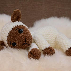 May include: A white crocheted lamb with brown fur accents on the face, ears, and feet. The lamb is lying on a white fluffy surface.