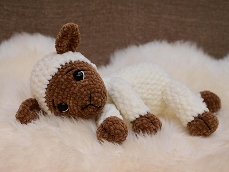 May include: A white crocheted lamb with brown fur accents on the face, ears, and feet. The lamb is lying on a white fluffy surface.
