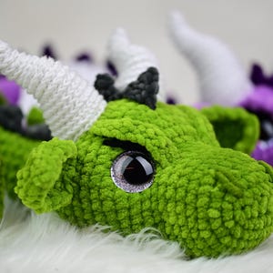 K&ouml;nnte beinhalten: Nahaufnahme eines gr&uuml;nen geh&auml;kelten Drachenkopfes mit wei&szlig;en H&ouml;rnern und gro&szlig;en, glitzernden Augen. Der Drache hat ein strukturiertes, pl&uuml;schiges Aussehen. Weitere Drachenk&ouml;pfe in Lila und Gr&uuml;n sind im Hintergrund zu sehen.