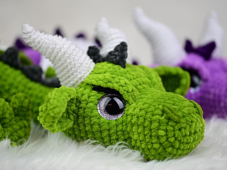 K&ouml;nnte beinhalten: Nahaufnahme eines gr&uuml;nen geh&auml;kelten Drachenkopfes mit wei&szlig;en H&ouml;rnern und gro&szlig;en, glitzernden Augen. Der Drache hat ein strukturiertes, pl&uuml;schiges Aussehen. Weitere Drachenk&ouml;pfe in Lila und Gr&uuml;n sind im Hintergrund zu sehen.