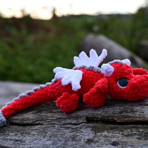 May include: A crocheted dragon plush toy featuring a vibrant red body, white wings, and a grey spine ridge that extends to a grey tail tip. The creature has large, dark, round eyes with white highlights and small grey horns on its head.