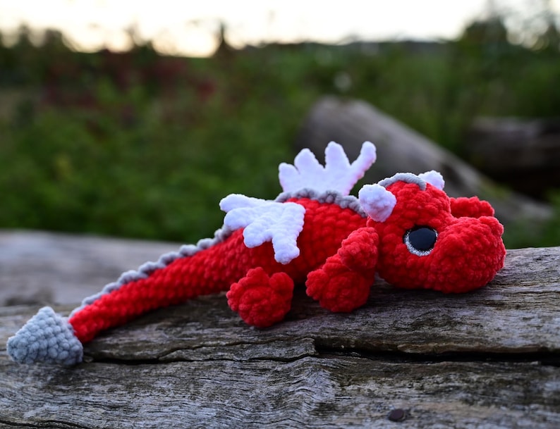 May include: A crocheted dragon plush toy featuring a vibrant red body, white wings, and a grey spine ridge that extends to a grey tail tip. The creature has large, dark, round eyes with white highlights and small grey horns on its head.