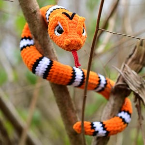 Pode incluir: Um brinquedo de cobra de croch&ecirc; laranja e preto com uma l&iacute;ngua vermelha. A cobra est&aacute; enrolada em torno de um galho.