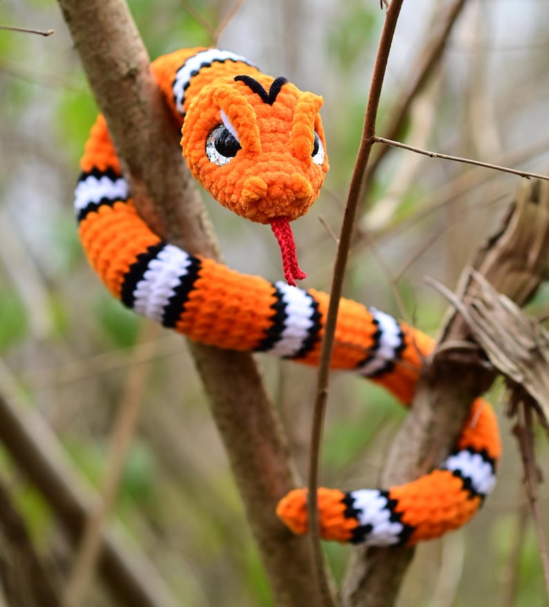 Pode incluir: Um brinquedo de cobra de croch&ecirc; laranja e preto com uma l&iacute;ngua vermelha. A cobra est&aacute; enrolada em torno de um galho.