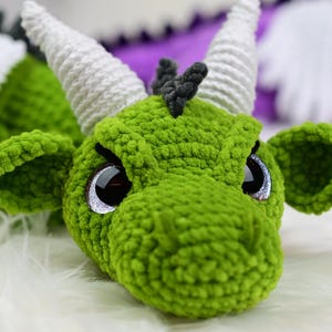 K&ouml;nnte beinhalten: Nahaufnahme eines handgefertigten gr&uuml;nen H&auml;keldrachens mit wei&szlig;en H&ouml;rnern und gro&szlig;en, reflektierenden Augen. Der Drache hat ein strukturiertes, pl&uuml;schiges Aussehen, mit kleinen schwarzen H&ouml;rnern und spitzen Ohren. Der Hintergrund ist unscharf.