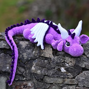 K&ouml;nnte beinhalten: Handgeh&auml;kelter Drache in Lila mit wei&szlig;en Fl&uuml;geln und H&ouml;rnern, auf einer Steinmauer liegend. Der Drache hat einen langen Schwanz und ist ein Fantasy-Artikel. Das Spielzeug ist aus weichem Garn gefertigt.