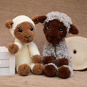 May include: Two crocheted sheep plushies, one white with brown accents and one brown with grey fur. The sheep are sitting on a brown surface.