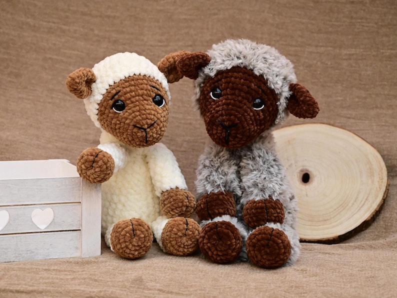 May include: Two crocheted sheep plushies, one white with brown accents and one brown with grey fur. The sheep are sitting on a brown surface.