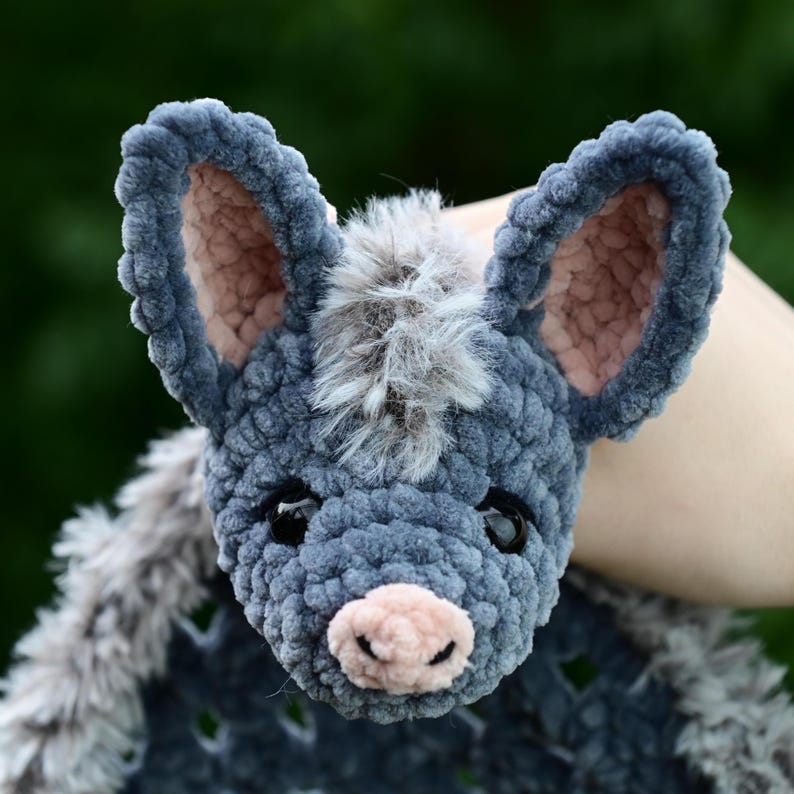 May include: A handmade, plush donkey head in shades of gray and pink. The donkey has large, floppy ears with pink inner ears, a pink nose, and black eyes. The donkey's mane is made of soft, gray fur.