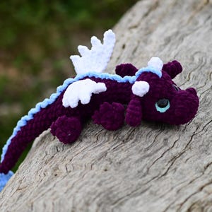 May include: A crocheted dragon plush toy featuring a deep purple body, white wings, and a light blue spine ridge. The creature has large, dark eyes with light blue irises, small horns on its head, and a long tail that ends in a light blue tip.